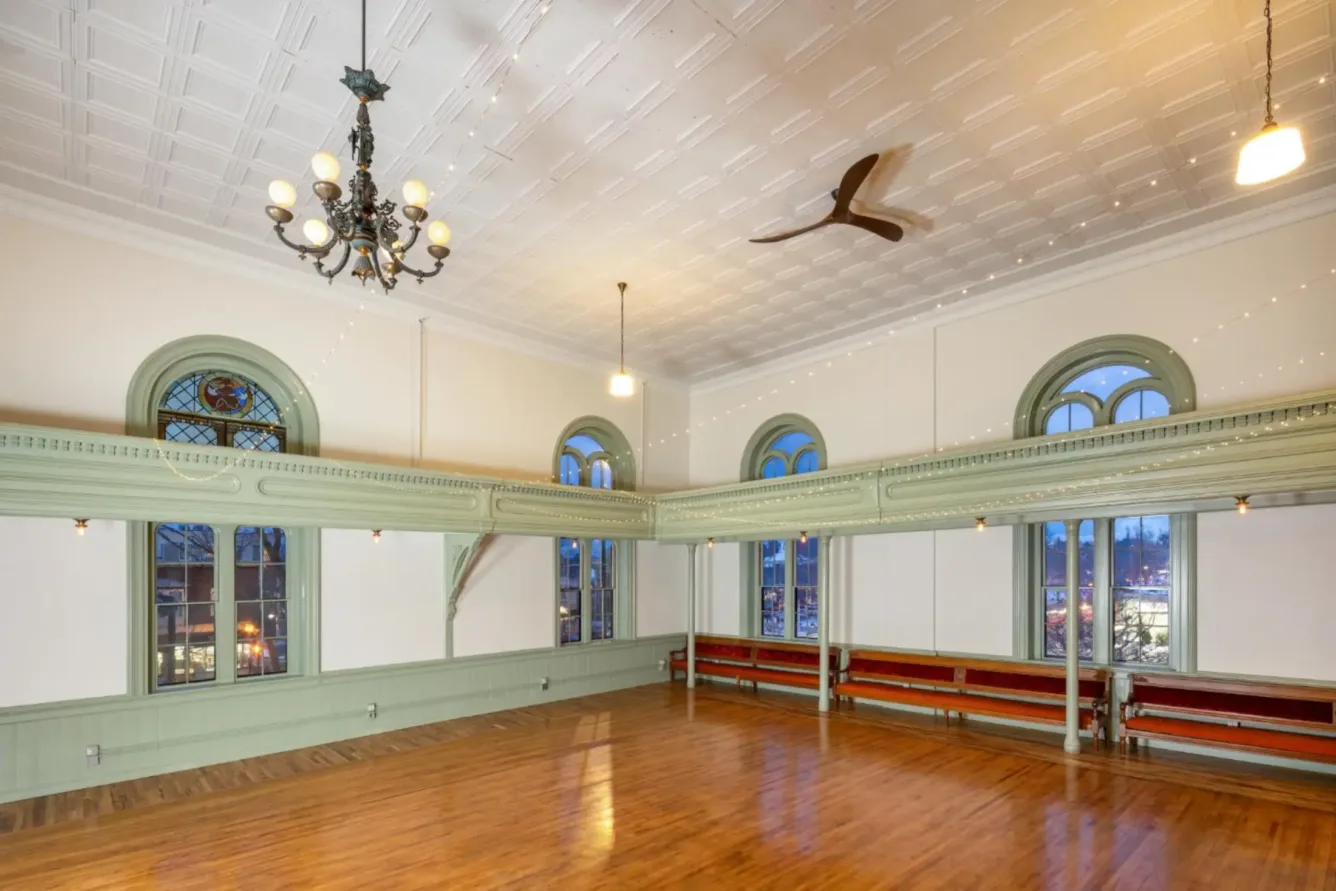 Spacious room with wooden floor, pale green trim, large arched windows, chandelier, ceiling fan, and red benches along the walls.