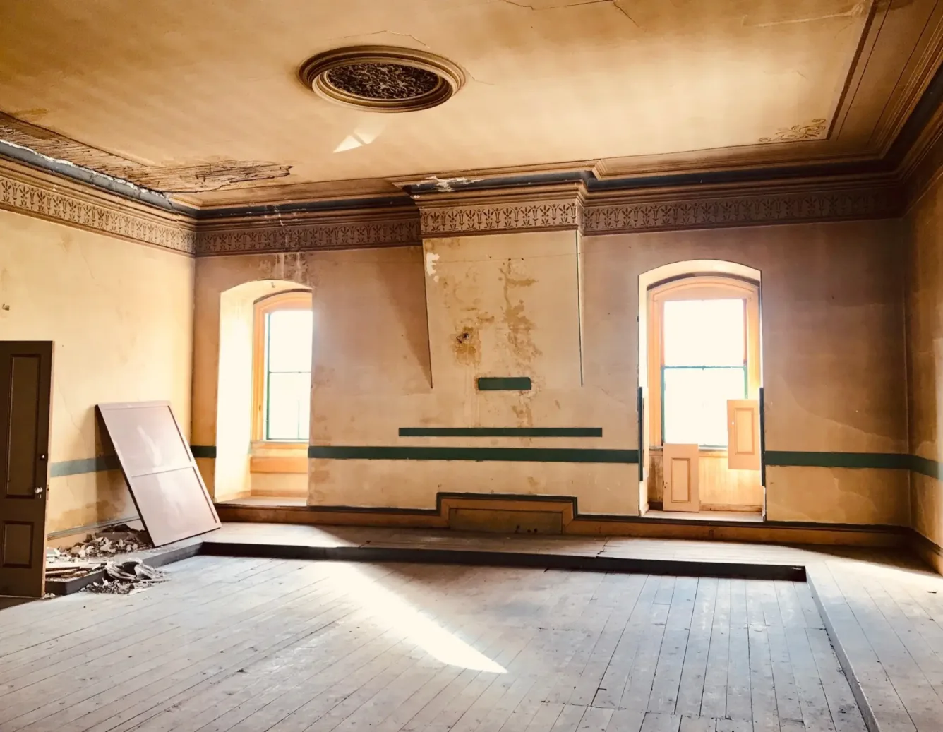Bright sunlight enters an old, empty room with peeling paint, wooden floors, arched windows, double doors, and ceiling moldings.