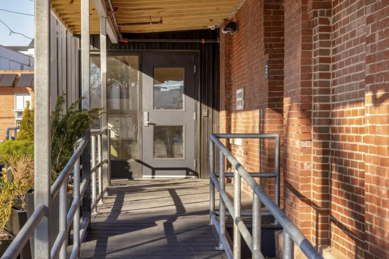A wooden ramp with metal railings leads to a black door set in a brick building under a partial wooden awning on a sunny day.