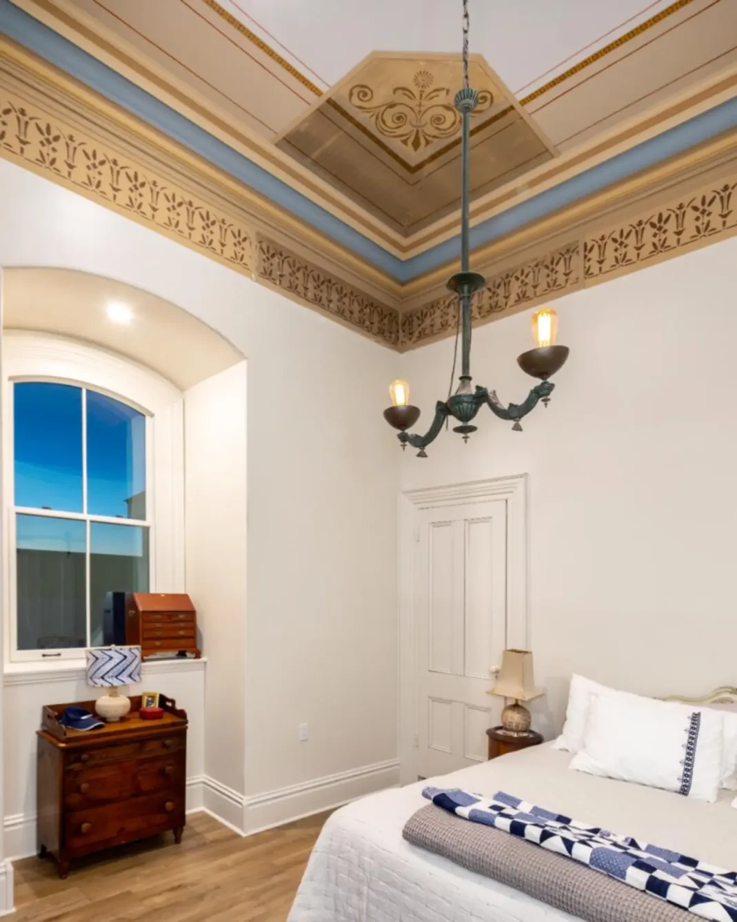 A bedroom with high ornate ceilings, a vintage chandelier, a bed with white linens, a wooden nightstand, and a dresser by a tall arched window at dusk.