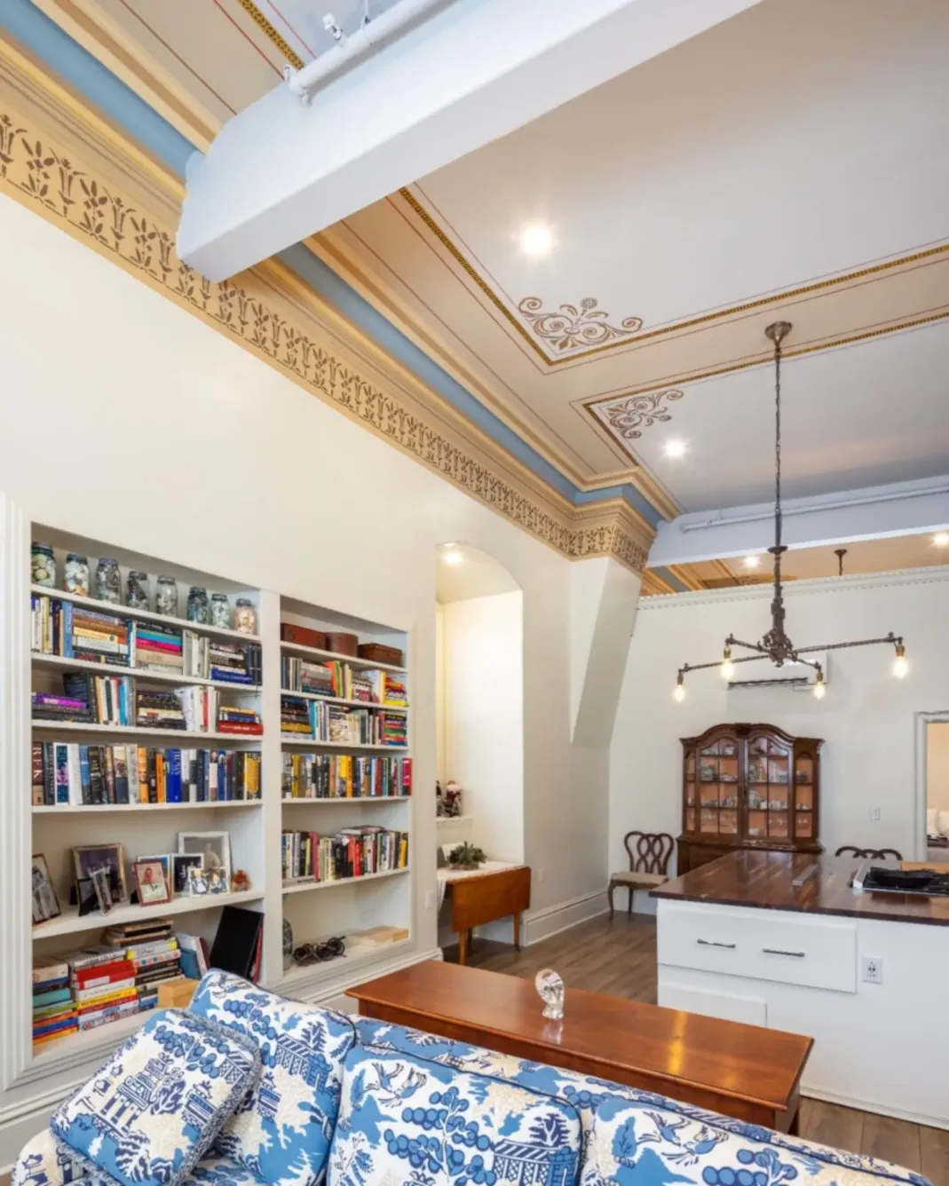 A living room with ornate ceiling molding, built-in bookshelves, a floral sofa, and a kitchen area with a chandelier and wooden dining furniture.