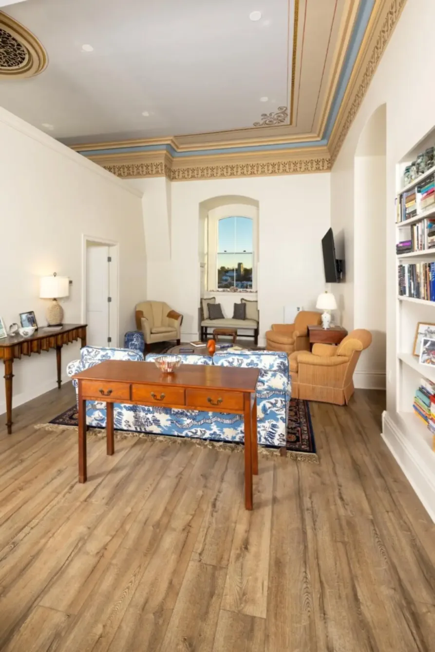 A bright living room with wood floors, a blue-patterned sofa, tan armchairs, wall shelves full of books, and a window showing a cityscape. Ornate ceiling molding and a desk are visible.