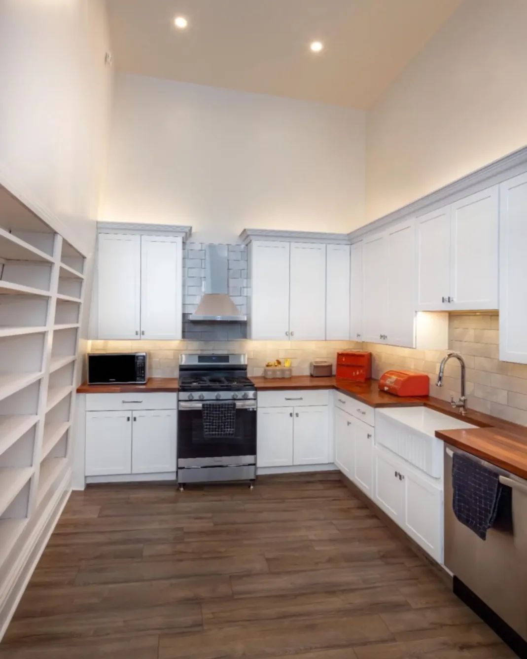 Modern kitchen with white cabinets, wood countertops, stainless steel appliances, open shelves on the left, and tile backsplash under bright ceiling lights.