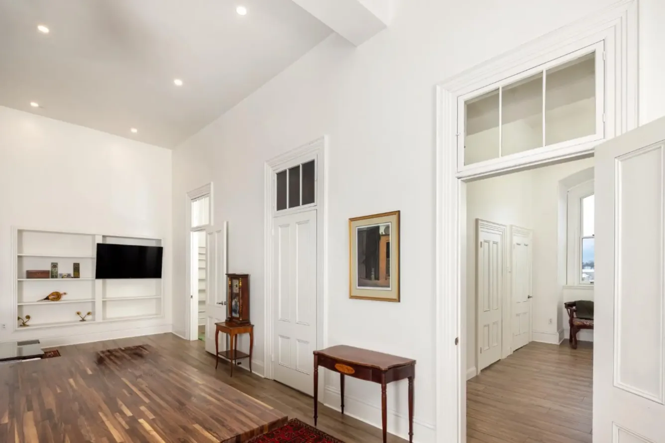 Spacious room with white walls, wood floors, built-in shelves, a TV, and several doors. A small table and framed art decorate the minimalist space.