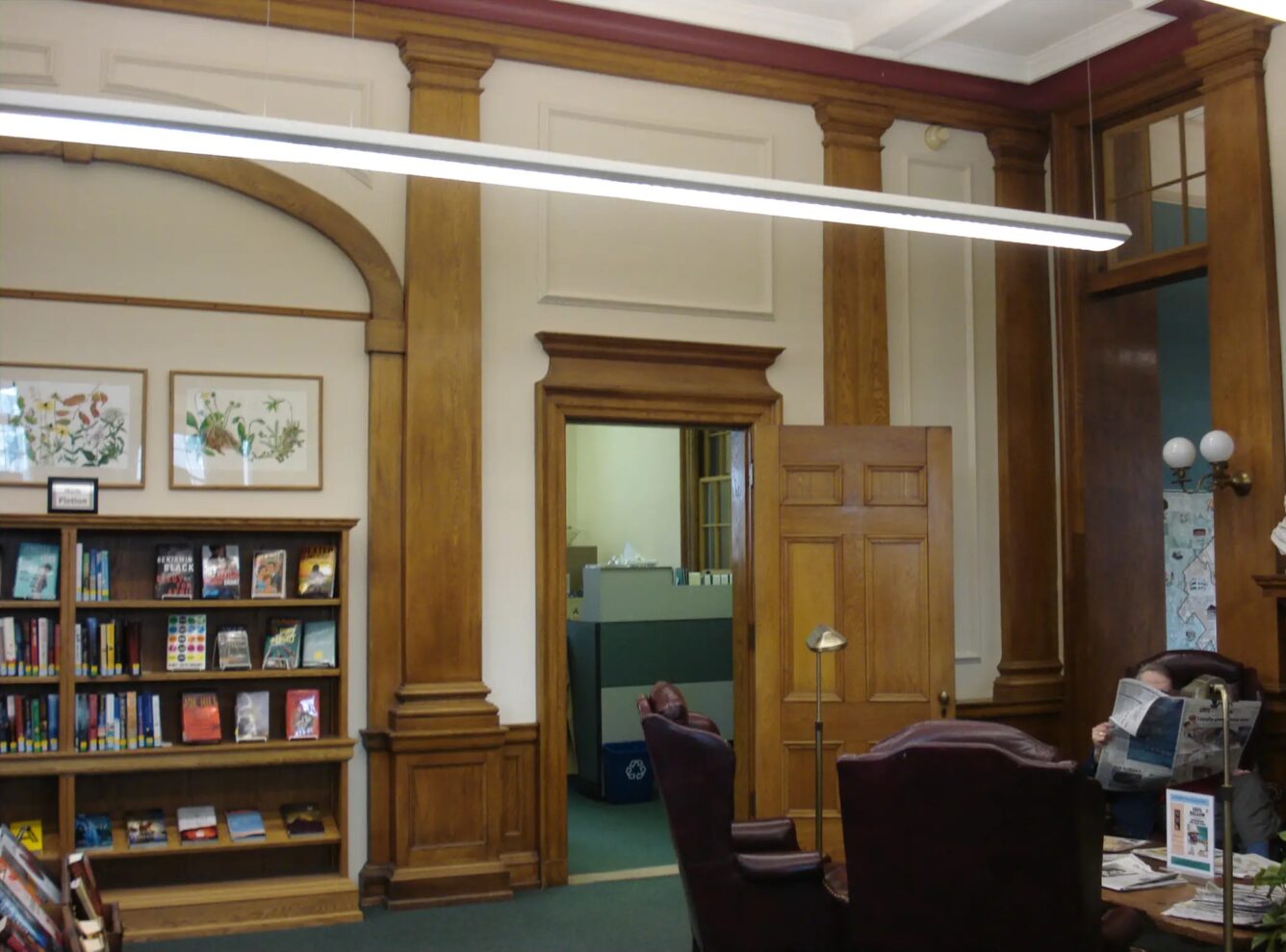 A traditional library reading room with wood paneling, bookshelves, archways, a desk chair, and an open door showing an adjacent office space.