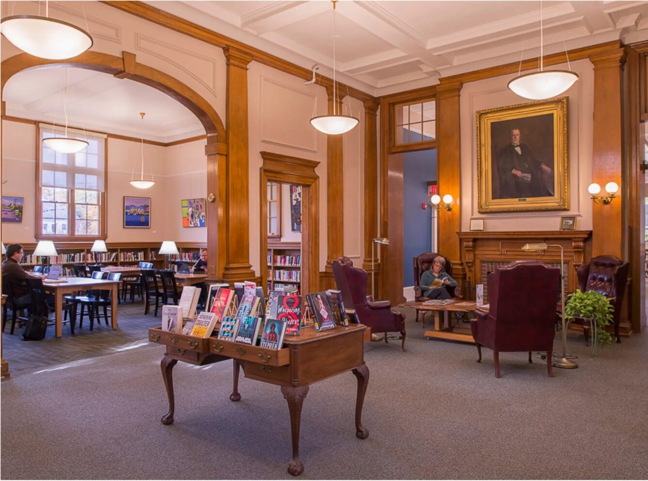 A library reading room with bookshelves, a display table with books, armchairs, large windows, and a portrait above a fireplace. People are reading and studying in the quiet space.