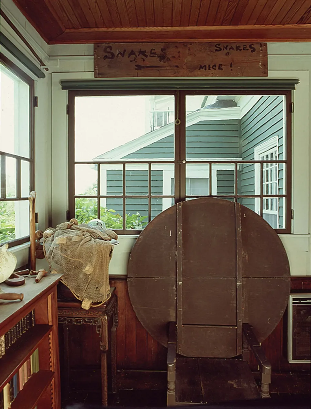 A wooden room featuring a hand-painted sign that reads SNAKES and MICE!, with a folded wooden table leaning against the wall and a fishing net sitting on a small table.