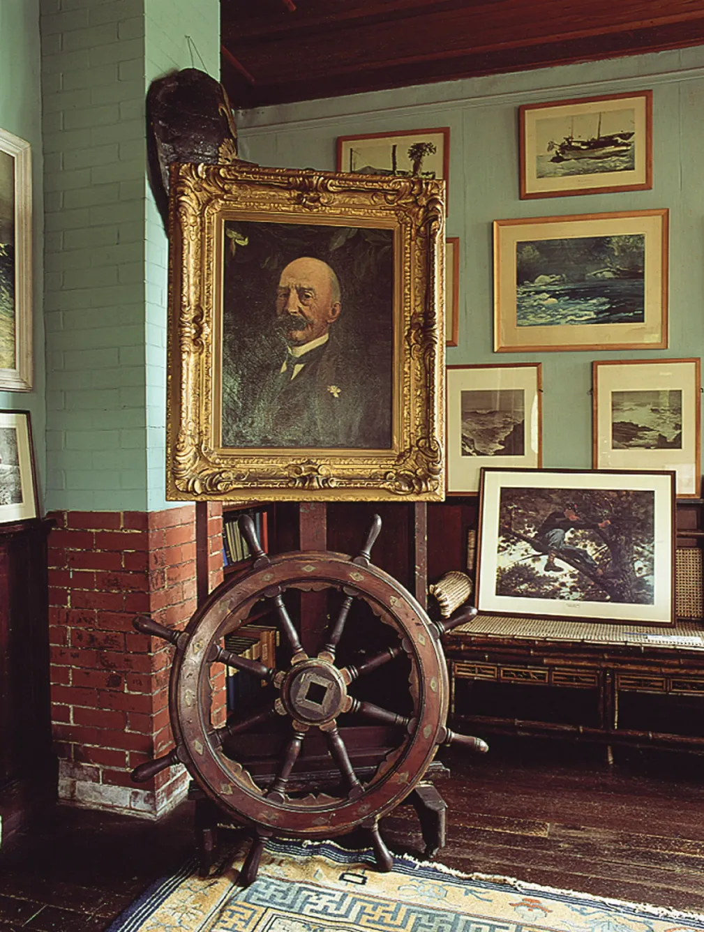 A wooden ships wheel standing in front of a framed portrait of a man, surrounded by framed paintings and sketches on a wall.