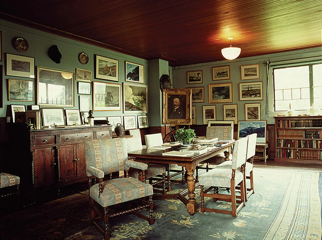 The studio's main room with a wood plank ceiling, walls hung with framed paintings, a dining table, and upholstered chairs.