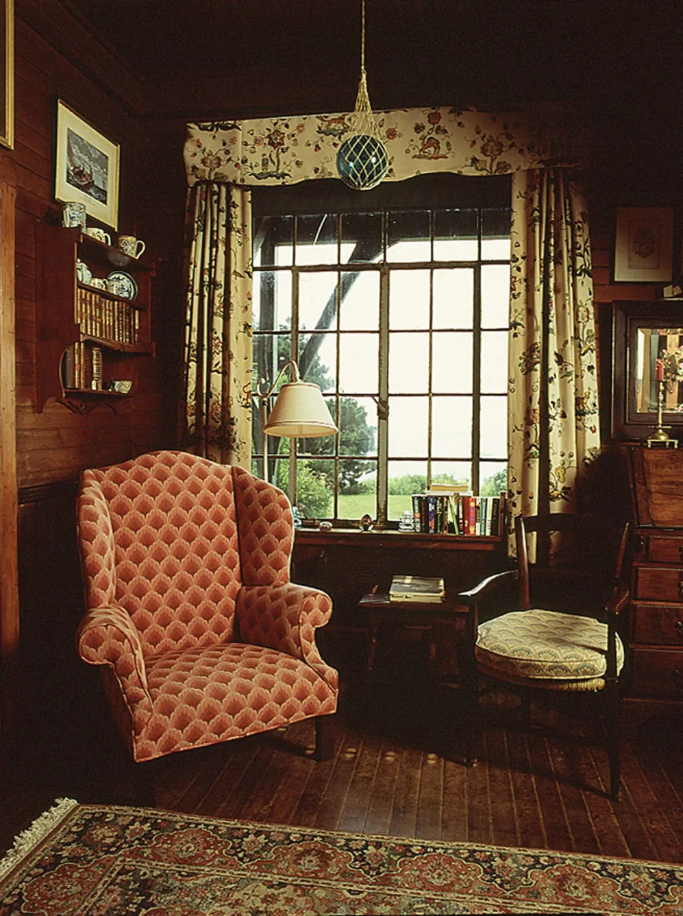 A corner of the studio with a red patterned wingback chair beside a window framed by floral curtains.