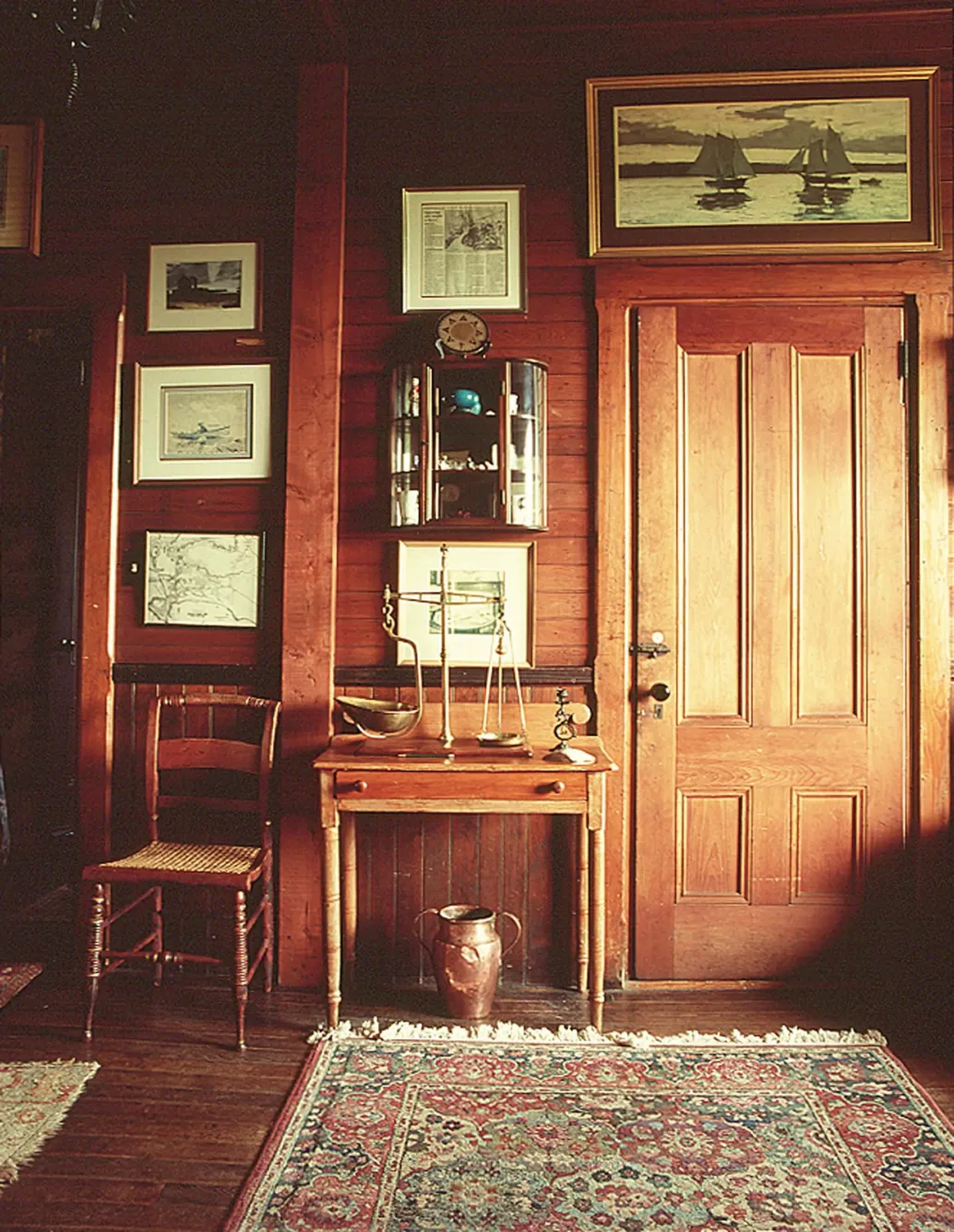 A wood-paneled room with framed nautical art, a small writing table holding a brass scale, a wooden chair, and a patterned rug on the floor.