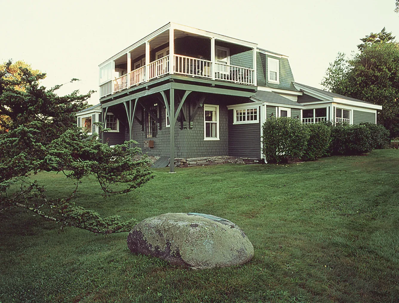 A two-story grey shingled house with a large wrap-around balcony, set in a grassy yard with a large boulder in the foreground.