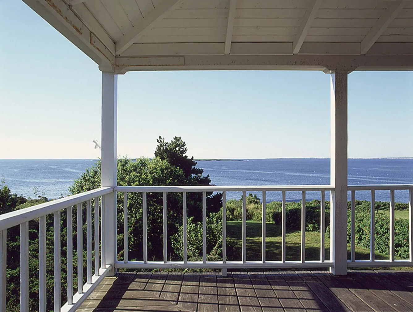 A view of the ocean and distant shoreline from a wooden porch with white railings.