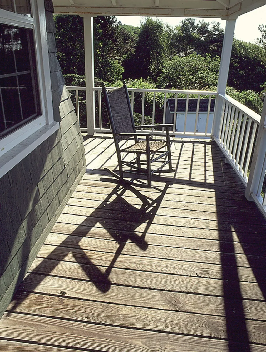 A rocking chair sits on a wooden porch, casting a long shadow across the floorboards.