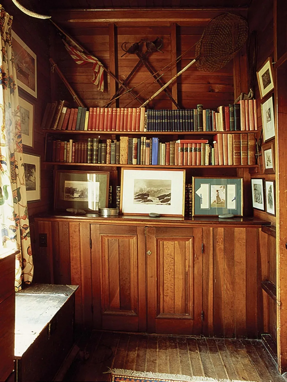 A wood-paneled room featuring two shelves of books, framed artwork, and vintage sports equipment mounted on the wall above storage cabinets.
