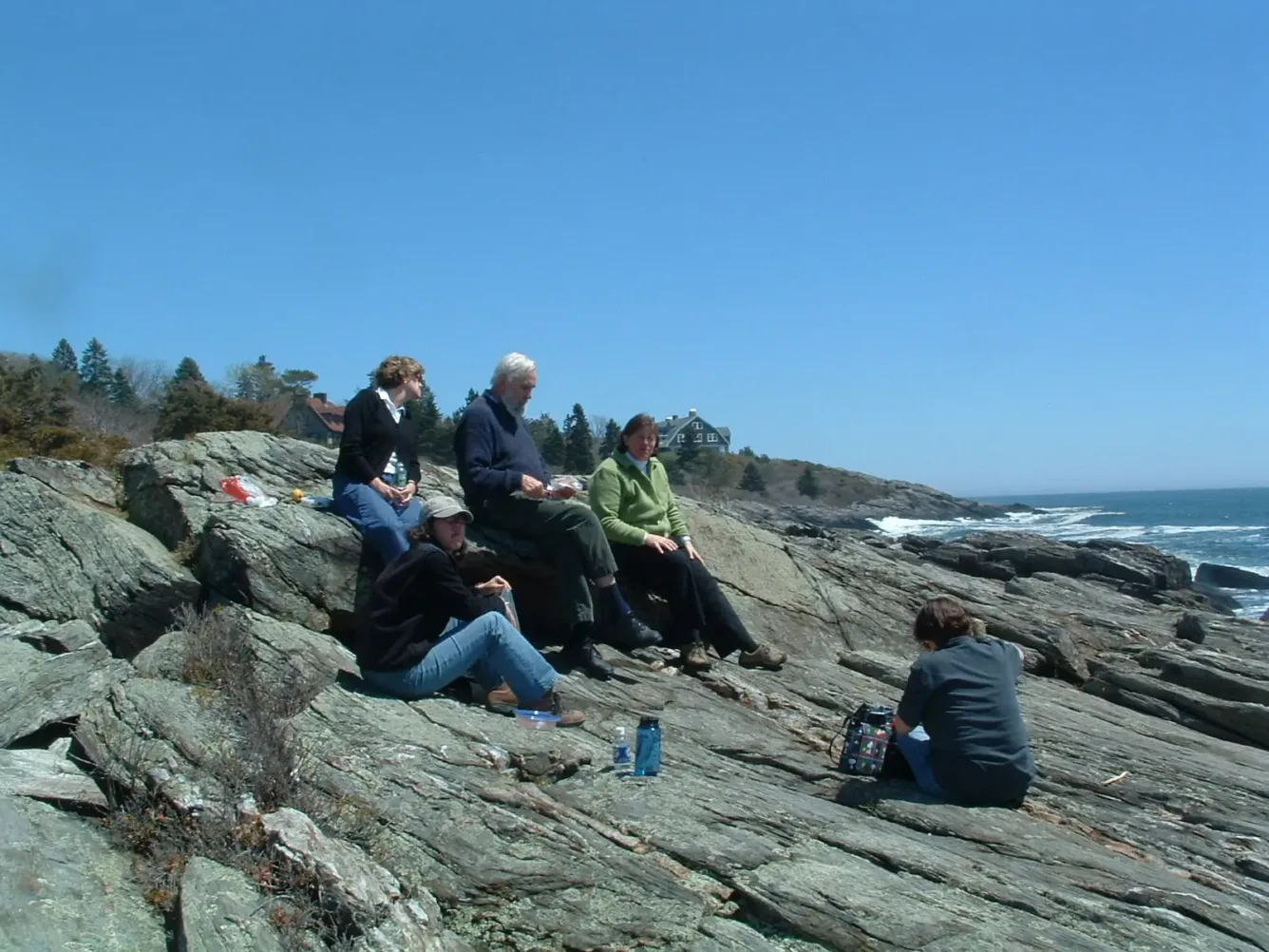 Members of the project team sit on the rocky shoreline below the studio, with the ocean behind them.