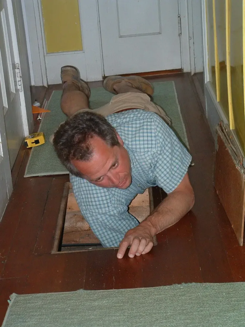 A man lies on a wood floor and looks into an open floor hatch during a survey of the studio.