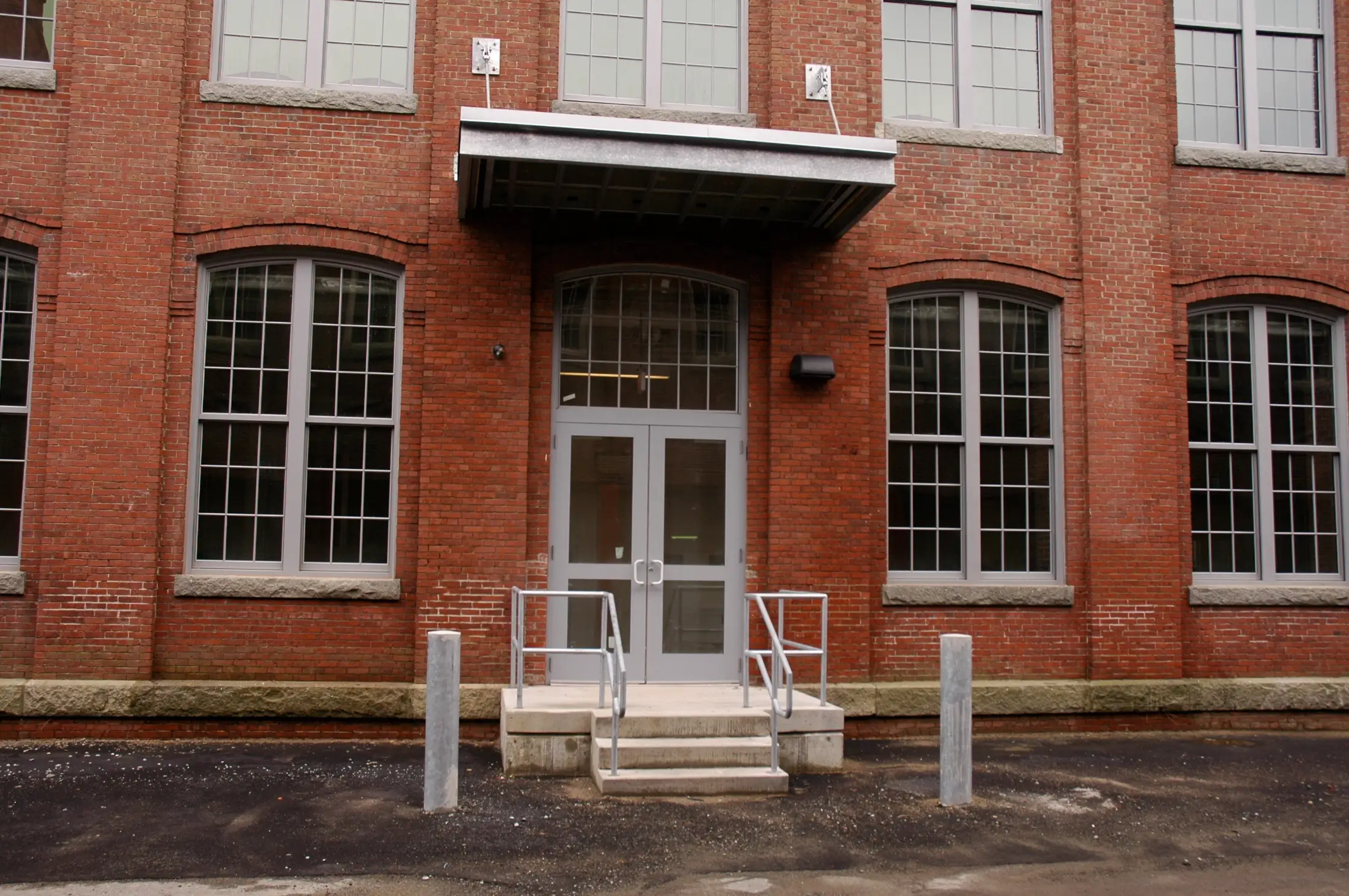 Exterior entrance of a brick industrial building with a metal awning over double glass doors and concrete steps with metal railings.
