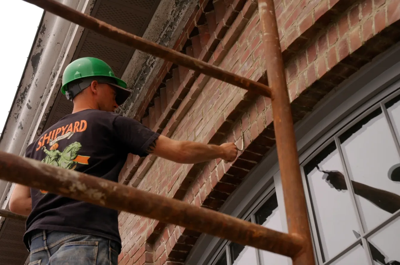 A construction worker wearing a green hard hat stands on scaffolding and repoints a brick wall above a window.