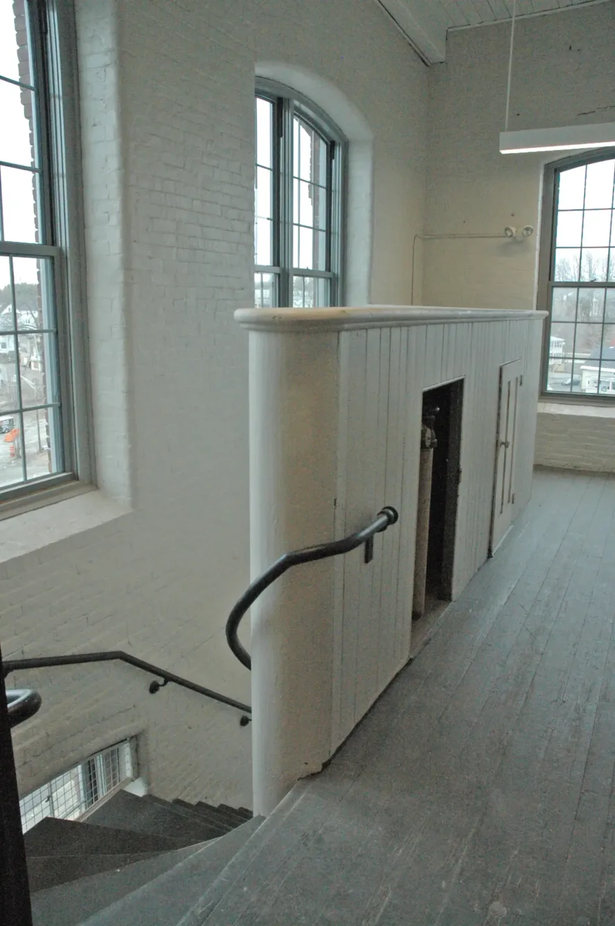 A wooden landing with a staircase leading downstairs, enclosed by a white vertical-panel railing and large industrial windows.
