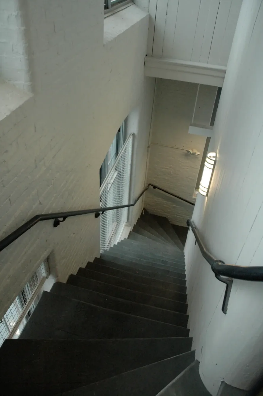 A downward view of a winding staircase with dark steps, white brick walls, and black handrails.