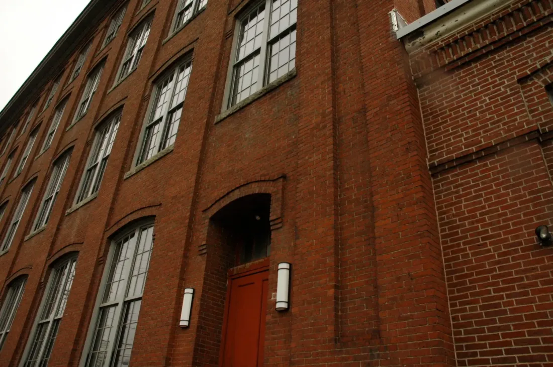 Low-angle view of a red brick industrial building with a recessed doorway and grid-patterned windows.