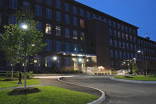A large, multi-story brick building with many windows at twilight, with exterior lights illuminating the entrance and a curved driveway in the foreground.