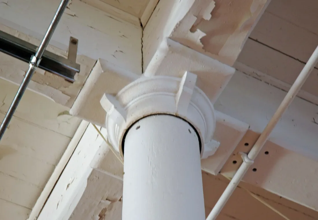 A white column capital supporting a ceiling beam with peeling paint.