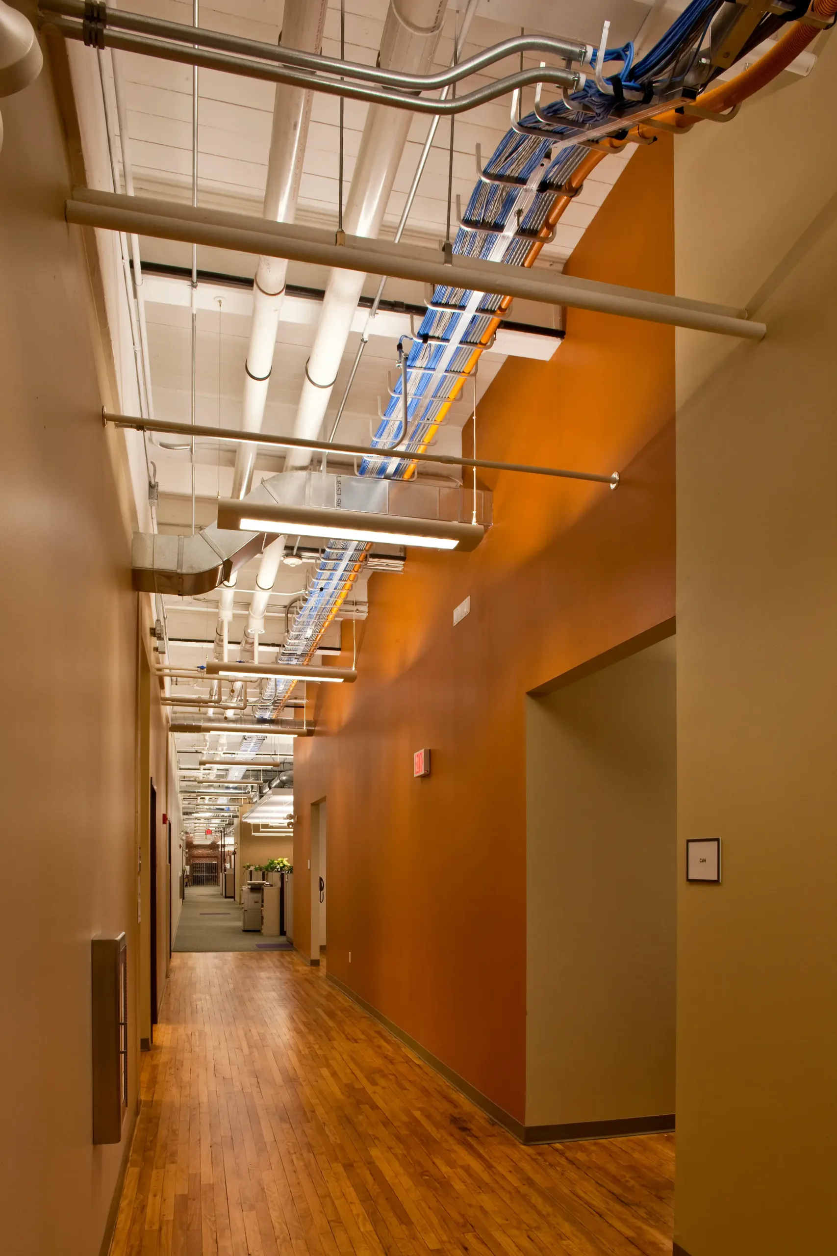 Long office corridor with orange accent walls, wood floors, and exposed ductwork and cable trays overhead.