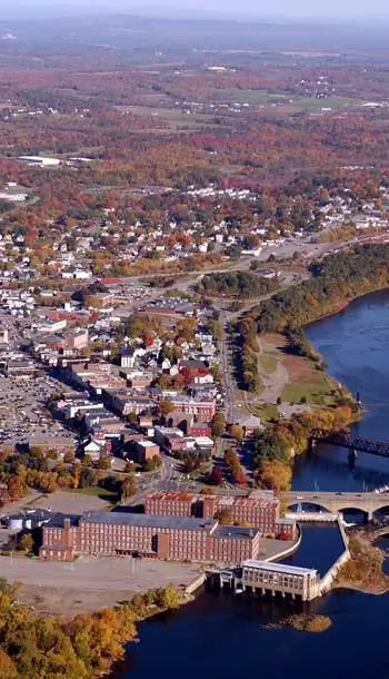 Aerial view of a town situated along a river, featuring a large brick industrial building and bridges, surrounded by autumn forests.