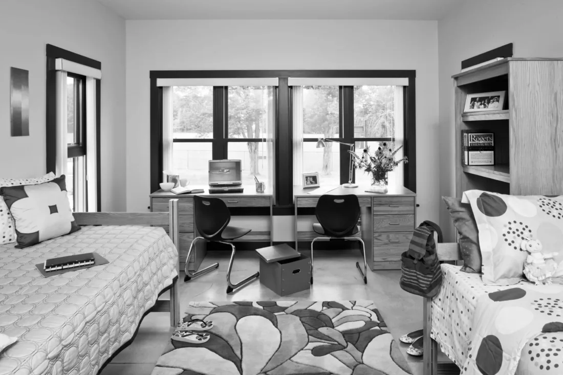 A shared dorm room with two beds, two desks with chairs in front of a window, and a patterned rug on the floor.