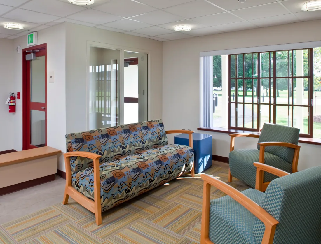 A waiting area with a patterned sofa, two green armchairs, and a large window looking out onto trees.