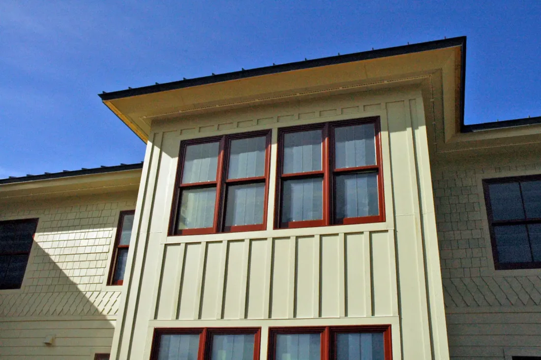 Exterior view of a light-colored house featuring dark red window frames and vertical siding.