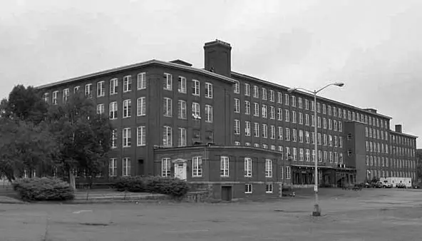 A large, multi-story brick industrial building with many windows sits behind a paved parking lot.