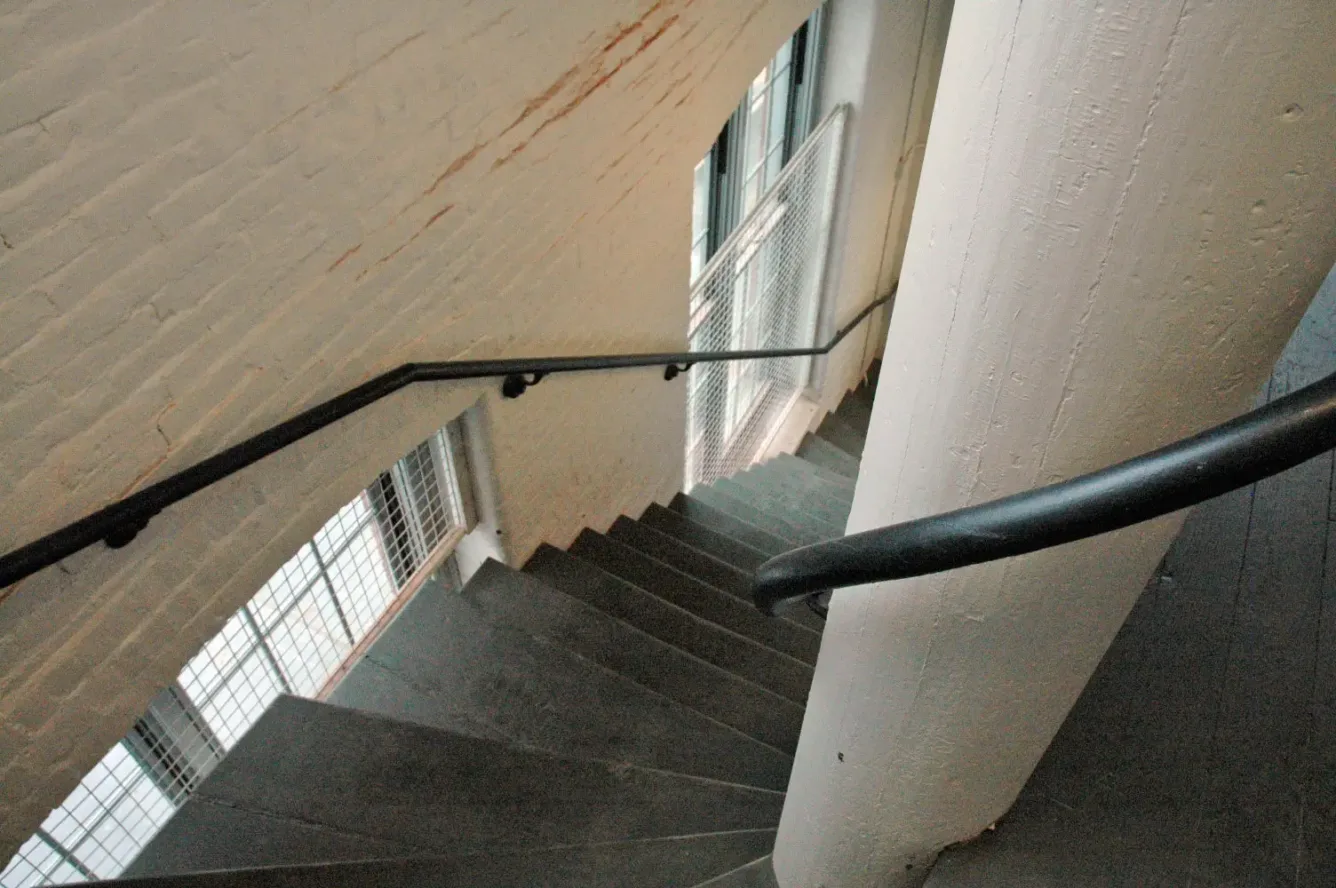 A winding stone staircase with a black metal handrail descends along a painted brick wall and a large structural column.