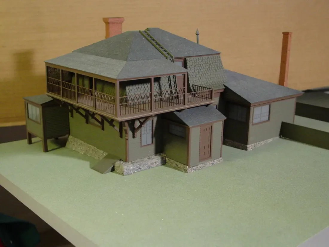 A scale model of a multi-level house with a second-story wraparound porch, two chimneys, and stone foundations.